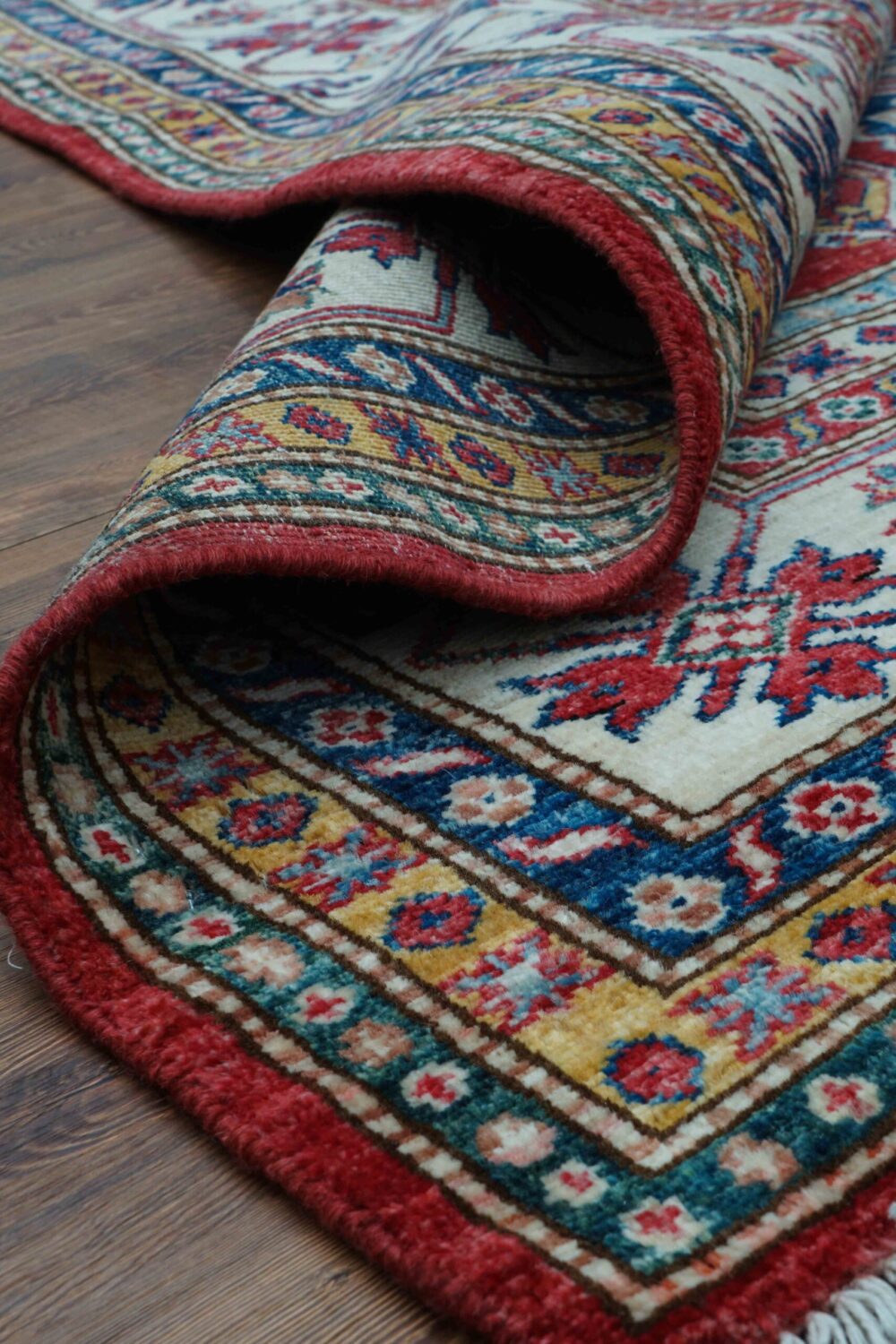 Super kazzak 6 x 8 rich red rug featuring intricate white and blue geometric medallions, surrounded by multicolored floral borders on a wooden floor.