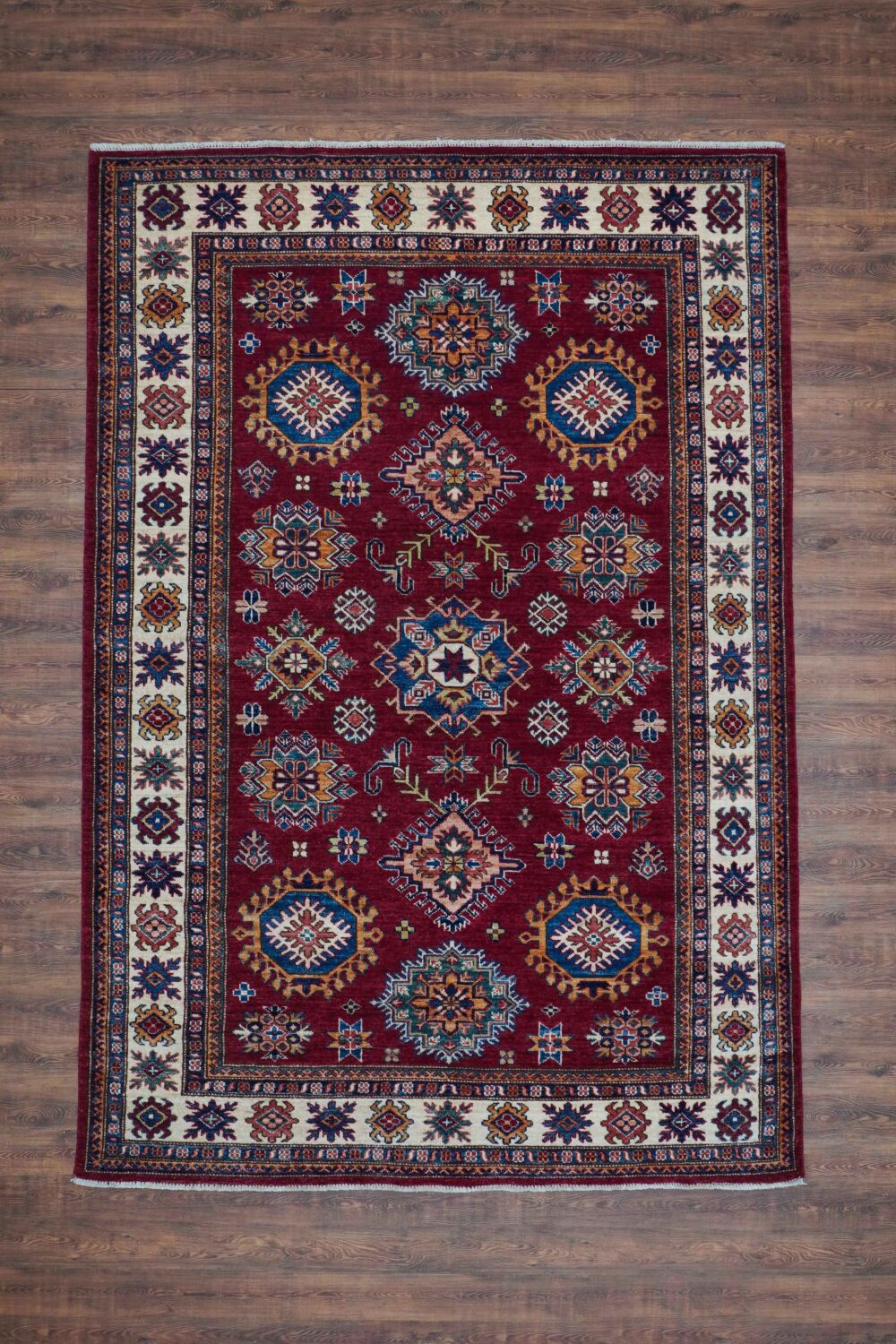 Super kazzak 5 x 7 rug showcases a deep red base adorned with vibrant floral and geometric motifs in gold, blue, and cream tones. The intricate border features complementary patterns, enhancing the overall richness and warmth, making it a perfect addition to traditional or eclectic interiors."