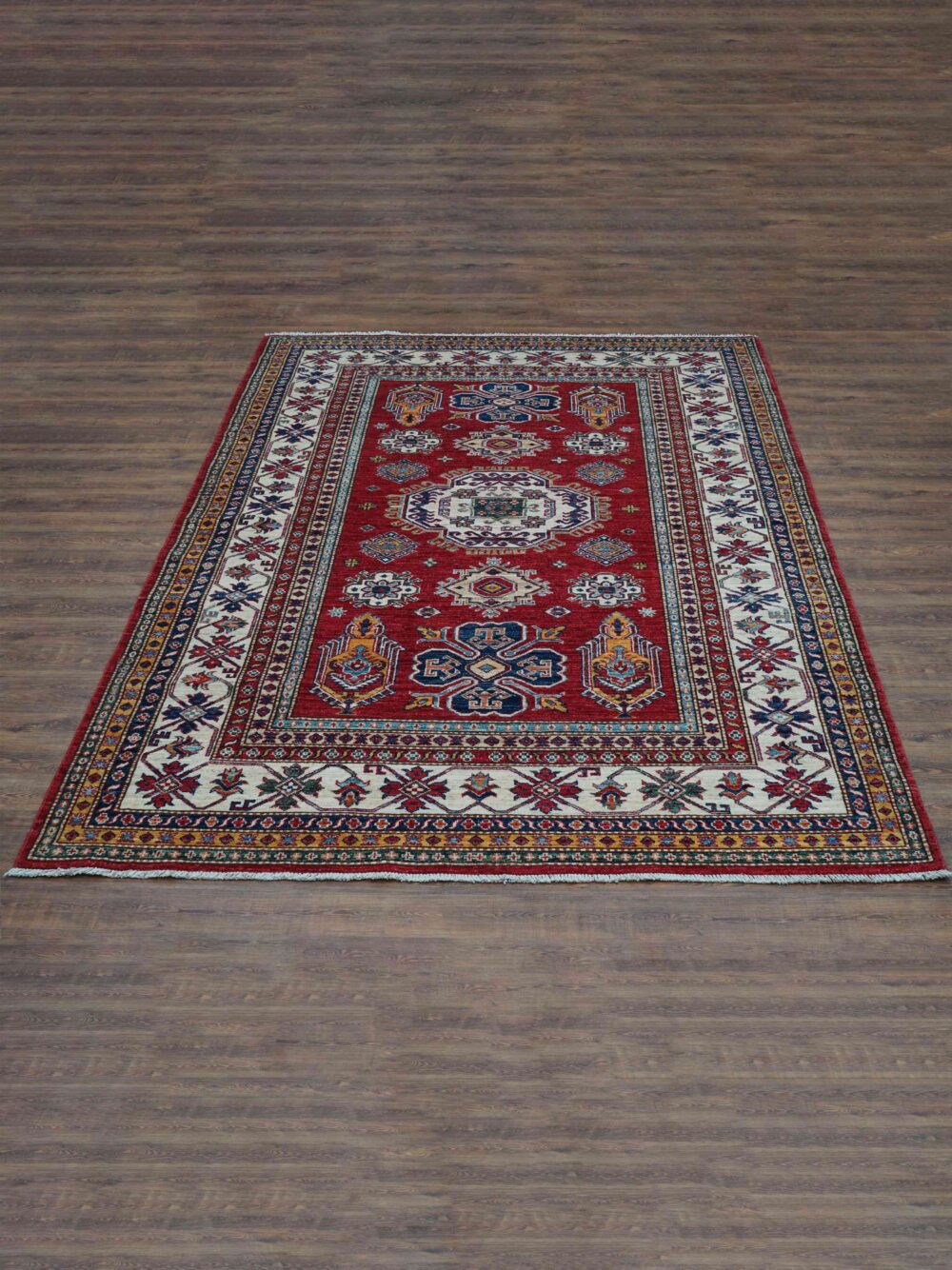 Super kazzak 6 x 8 rich red rug featuring intricate white and blue geometric medallions, surrounded by multicolored floral borders on a wooden floor.